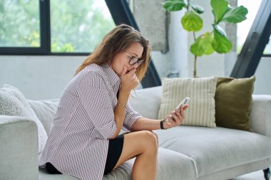 Üzgün, ciddi, orta yaşlı ve akıllı telefonlu bir kadın kanepesinde oturuyor. Kötü haberler, iş hayatında sorunlar, vücut ağrısı, akıl sağlığı konsepti