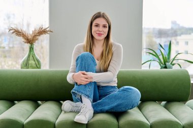 Evdeki kanepede oturan rahat ve güzel genç bir kadının portresi. Çekici bir kadın oturma odasındaki kanepede dinlenirken kameraya bakıyor. Boş zaman mutluluğu rahat yaşam tarzı gençlik konsepti
