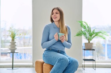 Rahatlamış genç bir kadının portresi, evinde elinde bardakla sandalyede oturuyor. Gülümseyen çekici bir kadın oturma odasında kupayla koltukta dinleniyor. Boş zaman mutluluğu rahat yaşam tarzı gençlik konsepti