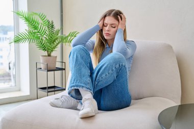 Evdeki kanepede oturan mutsuz, üzgün genç bir kadın, yüzü yakın çekim. Duygusal fiziksel acı çeken üzgün bir kadın. Sorunlar ruhun ve bedenin sağlığı, gençlik kavramı