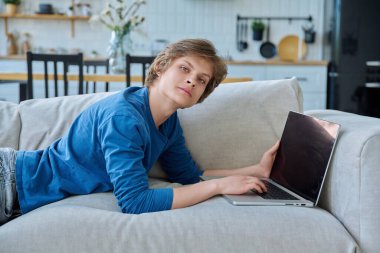 Evde dizüstü bilgisayarıyla kanepede uzanmış kameraya bakan yakışıklı bir delikanlı. Gençlik yaşı, ergenlik, yaşam tarzı, teknoloji konsepti