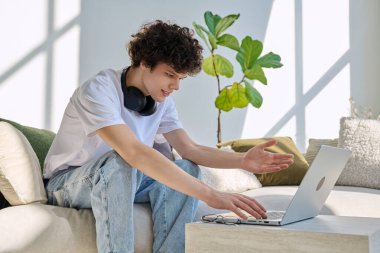 Evdeki kanepede dizüstü bilgisayar kullanan yakışıklı bir erkek portresi. 20 yaşında bilgisayar başında yazı yazan kıvırcık bir öğrenci. Yaşam tarzı, eğitim, teknoloji, gençlik kavramı