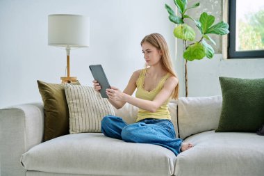 Genç ve güzel bir kız öğrenci dijital tabletle online sohbet konferansı yapıyor. Evde, kanepede oturuyor. İletişim öğrenme eğitimi için dijital teknolojiler boş zamanlarında blog yazarlığı