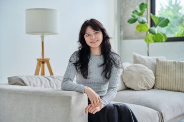 Evdeki kanepede oturan rahat ve güzel genç bir kadının portresi. Oturma odasında kameraya bakan çekici bir kadın. Boş zaman mutluluğu rahat yaşam tarzı gençlik konsepti