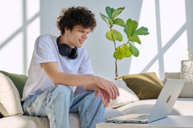 Genç yakışıklı kıvırcık adam internette sohbet etmek için bilgisayarını kullanıyor. Evde, koltukta oturuyor. İletişim öğrenimi için dijital teknolojiler boş zamanlarında sosyal medya bloglarında çalışır