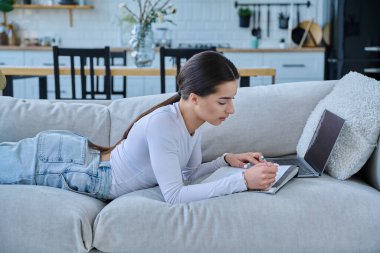 Evde dizüstü bilgisayarla kanepede uzanmış sınav için notlar alan genç üniversiteli kız. Eğitim e-öğrenim kursu online ders yaşam tarzı gençlik kavramı