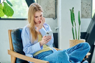 Üzgün, ağlayan, akıllı telefona bakan, evdeki sandalyede oturan genç bir kadın. İletişim kurmak için telefon kullanan bir kadın, eğitim için mobil uygulamalar sosyal ağlar