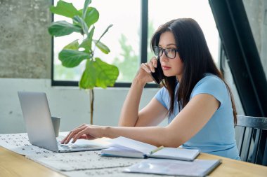 Bilgisayarlı dizüstü bilgisayarla çalışan, akıllı telefondan konuşan, mutfaktaki masada oturan genç bir kadın. Teknoloji, iş, iş, 30 'ların insan kavramı