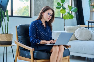 Bilgisayarda yazı yazan, sandalyeye oturan, bilgisayar ekranına bakan orta yaşlı güzel bir kadının portresi. Teknoloji, yaşam tarzı, iş, eğlence, insan kavramı