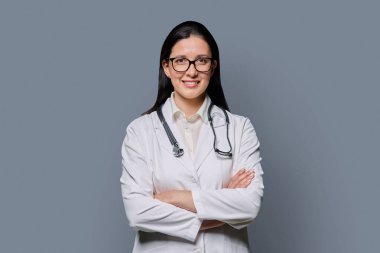 Gri stüdyo arka planında steteskobu olan, beyaz laboratuvar önlüklü, kendine güvenen kadın doktor portresi. Tıp, insanlar, sağlık personeli, sağlık hizmetleri kavramı