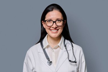 Gri stüdyo arka planında steteskobu olan beyaz laboratuvar önlüklü gülümseyen kadın doktor portresi. Tıp, insanlar, sağlık personeli, sağlık hizmetleri kavramı