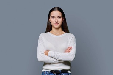 Kollarını kavuşturup gri stüdyonun arka planında kameraya bakan kendine güveni tam genç bir kız. Başarılı genç kız öğrenci üniversite yaşı gençlik için reklam ürünleri ve hizmetleri