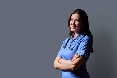 Özgüvenli orta yaşlı kadın doktor portresi. Mavi üniformalı, steteskoplu, gri stüdyo arka planında çapraz kollu hemşire. Kopya odası. Tıp, insanlar, sağlık personeli, sağlık hizmetleri kavramı