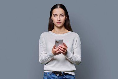 Gri stüdyo arka planında akıllı telefon tutan ciddi bir genç kızın portresi. Genç bayan cep telefonu kullanıyor, kameraya bakıyor. Teknoloji, eğitim, eğlence ve iletişim için mobil uygulamalar