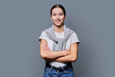 Kollarını kavuşturup gri stüdyonun arka planında kameraya bakan kendine güveni tam genç bir kız. Başarılı gülümseyen genç kız üniversite öğrencisi. Gençler için reklam ürünleri ve hizmetler.