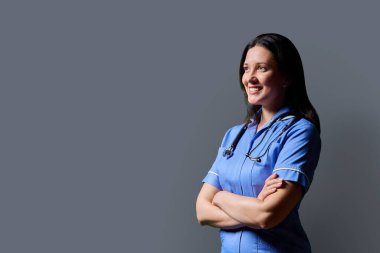 Özgüvenli orta yaşlı kadın doktor portresi. Mavi üniformalı, steteskoplu, gri stüdyo arka planında çapraz kollu hemşire. Kopya odası. Tıp, insanlar, sağlık personeli, sağlık hizmetleri kavramı