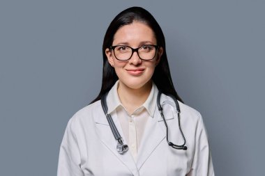 Gri stüdyo arka planında steteskobu olan beyaz laboratuvar önlüklü gülümseyen kadın doktor portresi. Tıp, insanlar, sağlık personeli, sağlık hizmetleri kavramı