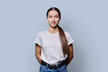 Gri stüdyo arka planında poz veren güzel, gülümseyen genç bir kadının portresi. Beyaz tişörtlü, kameraya bakan 18 yaşında başarılı bir kız. Güzellik yaşam tarzı sağlık gençlik kavramı