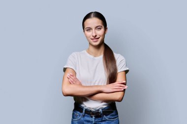 Kollarını kavuşturup gri arka planda poz veren güzel, gülümseyen genç bir kadının portresi. Beyaz tişörtlü, kameraya bakan kendinden emin bir genç kız. Eğitim yaşam tarzı sağlık gençleri