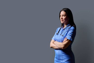 Özgüvenli orta yaşlı kadın doktor portresi. Mavi üniformalı, steteskoplu, gri stüdyo arka planında çapraz kollu hemşire. Kopya odası. Tıp, insanlar, sağlık personeli, sağlık hizmetleri kavramı