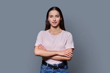 Kollarını kavuşturup gri stüdyonun arka planındaki kameraya bakan kendine güvenen genç kız. Başarılı genç kız öğrenci üniversite yaşı gençlik için reklam ürünleri ve hizmetleri