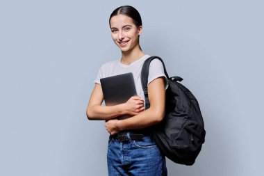 Sırt çantalı, dizüstü bilgisayarı gri arka planda kameraya bakan güzel, gülümseyen genç kız öğrencinin portresi. Eğitim, gençlik, öğrenme, teknoloji, üniversite insanları kavramı