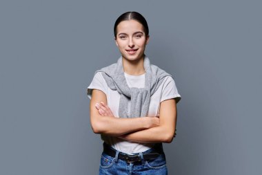 Kollarını kavuşturup gri stüdyonun arka planında kameraya bakan kendine güveni tam genç bir kız. Başarılı gülümseyen genç kız üniversite öğrencisi. Gençler için reklam ürünleri ve hizmetler.