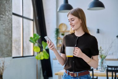 Elinde bir bardak içkiyle evde duran akıllı telefonu kullanan genç bir kadın. Mesajlaşan genç kız gezici uygulamaları okuyor. İnternet teknolojisi. Boş zaman iletişim yaşam tarzı eğitimi için internet hizmetleri.
