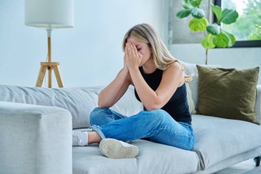 Mutsuz, üzgün, başı ağrıyan genç bir kadın, evdeki kanepede oturuyor. Duygusal fiziksel acı çeken üzgün bir kadın. Sorunlar ruhun ve bedenin sağlığı ve gençliğin sorunları