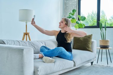 Video sohbet konferansı için akıllı telefonu kullanan genç bir kadın evde oturmuş web kamerasından, telefon ekranından konuşuyor. Eğitim teknolojileri eğlence iletişimi, gençlik kavramı