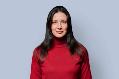 Kırk yaşında, kırmızı kazaklı, açık gri stüdyo arka planında poz veren güzel gülümseyen bir kadının portresi. Orta yaş, güzellik, sağlık ve yaşam tarzı kavramları.