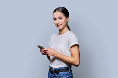 Gri arka planda akıllı telefonu tutan gülümseyen genç kızın portresi. Genç bir bayan telefon kullanıyor, kameraya bakıyor. Eğitim için gezici teknoloji uygulamaları boş zaman iletişimi uygulamaları