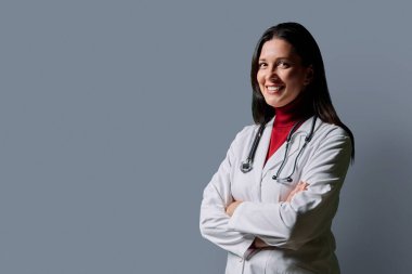 Beyaz laboratuvar önlüklü, gri stüdyo arka planında steteskobu olan gülümseyen, kendine güvenen kadın doktor portresi. Tıp, insanlar, sağlık personeli, sağlık hizmetleri kavramı