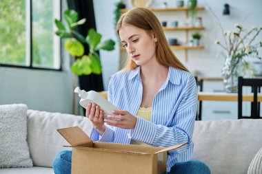 Genç, mutlu, karton kutuları internet siparişiyle açan, cilt bakımı parfümlü ev yapımı kimyasallar alan, evinde kanepede oturan bir kadın. E-alışveriş, e-market, teslimat servisi.