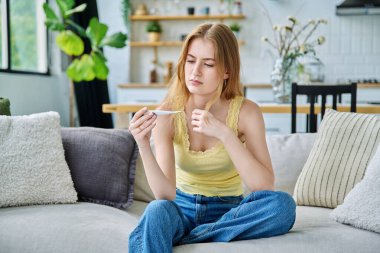 Evde oturan termometreye bakan üzgün genç kız. Yüksek sıcaklıkta mevsimsel viral hastalıklar, hastalık, sağlık gençliği, ergenlik kavramı