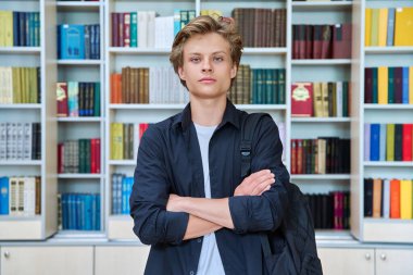 Kütüphanede kollarını kavuşturmuş ciddi bir genç öğrencinin portresi. Kendine güveni tam erkek, 15-16 yaşında. Okulun içindeki kameraya bakıyor. Eğitim bilgisi okulu ergenliği