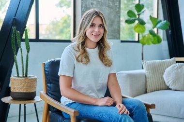 Rahatlamış genç bir kadının portresi sandalyede oturuyor. Çekici bir kadın oturma odasında dinlenirken kameraya bakıyor. Boş zaman mutluluğu rahat yaşam tarzı gençlik konsepti