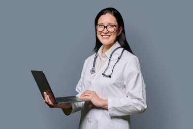 Gri arka planda dizüstü bilgisayarı olan kadın doktorun portresi. Gülümseyen orta yaşlı kadın doktor elinde bilgisayarla kameraya bakıyor. Tıp, e-tıp, teknoloji, sağlık konsepti