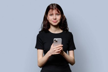 Akıllı telefonlu güzel, gülümseyen genç kız öğrencinin portresi gri stüdyo arka planındaki kameraya bakıyor. Eğitim, ergenlik, iletişim, teknoloji, lise insanları kavramı