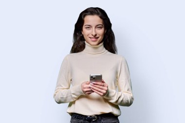 Beyaz stüdyo arka planında akıllı telefonu tutan gülümseyen genç kızın portresi. Genç bayan cep telefonu kullanıyor, kameraya bakıyor. Teknoloji, eğitim, eğlence ve iletişim için mobil uygulamalar