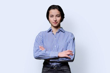 Beyaz arka planda kameraya bakan 18 yaşında güzel bir kızın portresi. Kollarını kavuşturmuş mavi gömlekli kendine güvenen bir üniversite öğrencisi. Güzellik sağlık eğitimi gençlik yaşam tarzı