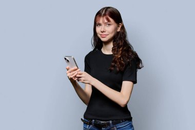 Akıllı telefonlu güzel, gülümseyen genç kız öğrencinin portresi gri stüdyo arka planındaki kameraya bakıyor. Eğitim, ergenlik, iletişim, teknoloji, lise insanları kavramı
