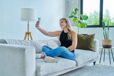 Genç, güler yüzlü, mutlu bir kadın akıllı telefon kullanarak selfie çekiyor. Oturma odasındaki kanepenin yanında oturuyor.
