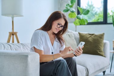 Orta yaşlı, rahat, gülümseyen mutlu bir kadın evde koltukta uzanmış akıllı telefon mesajları okuyor. Boş zaman iletişimi için mobil uygulamalar iş eğitimi teknoloji yaşam tarzı insanlar kavramı