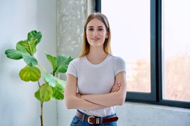 17-19 yaşlarındaki genç bir kızın portresi. Evde çapraz kollarla kameraya bakarak kendinden emin gülümsüyor. Uzun düz saçlı güzel sarışın kız. Gençlik yaşam tarzı güzellik moda konsepti