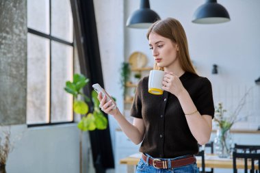Elinde bir bardak içkiyle evde duran akıllı telefonu kullanan genç bir kadın. Mesajlaşan genç kız gezici uygulamaları okuyor. İnternet teknolojisi. Boş zaman iletişim yaşam tarzı eğitimi için internet hizmetleri.
