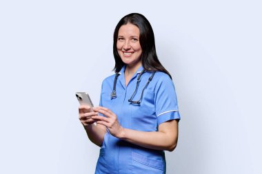 Beyaz stüdyo arka planında akıllı telefonlu gülümseyen kadın hemşire portresi. Mobil uygulamalar, tıbbi hizmetlerdeki teknolojiler, sağlık, profesyonel yardım, tıbbi bakım kavramı