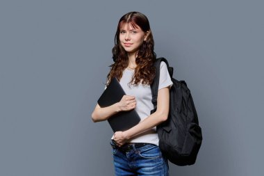 Sırt çantalı, dizüstü bilgisayarı gri arka planda kameraya bakan güzel, gülümseyen genç bir kızın portresi. Eğitim, okul, öğrenme, teknoloji, okul çocukları kavramı