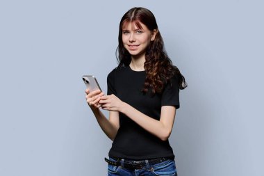 Akıllı telefonlu güzel, gülümseyen genç kız öğrencinin portresi gri stüdyo arka planındaki kameraya bakıyor. Eğitim, ergenlik, iletişim, teknoloji, lise insanları kavramı