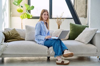 Genç bir kadının portresi kanepede oturuyor ve dizüstü bilgisayarda ev içinde daktilo yazıyor. Üniversite öğrencisi, internetten sosyal medya blogu alışverişini rahatlatmak için sınava hazırlanıyor.